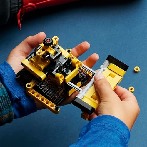 Child assembling a yellow LEGO bulldozer on a blue surface.