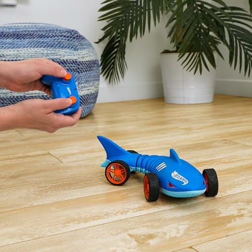 Hands holding remote controlling a blue shark-shaped toy car on wooden floor.